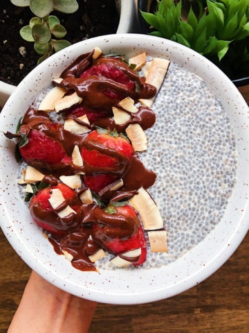 Hand holding white bowl with chia pudding in it as well as strawberries, coconut flakes, and drizzled with chocolate.