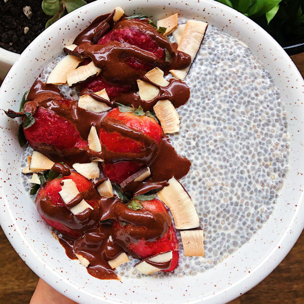 Hand holding white bowl with chia pudding in it as well as strawberries, coconut flakes, and drizzled with chocolate.