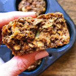 Hand holding carrot muffin cut in half with muffin tin of other carrot muffins in the background.