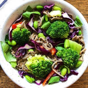 White bowl with buckwheat noodles, red cabbage, red bell pepper, edamame and broccoli with peanut lime sauce mixed in.