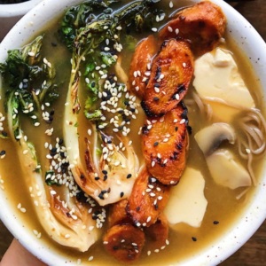 White bowl with miso broth with sliced bok choy, roasted carrots, soft tofu, mushrooms and buckwheat noodles with sesame seeds on top.