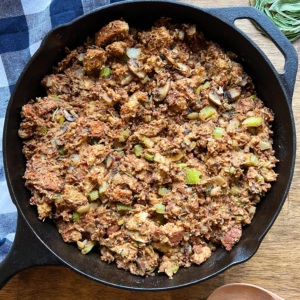 Wooden table with a blue and white cloth and a cast iron dish with lentil mushroom vegan stuffing inside and fresh sage as garnish.