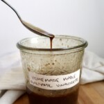Spoon resting on a glass jar filled with homemade maple balsamic vinaigrette, with a white cloth in the background.