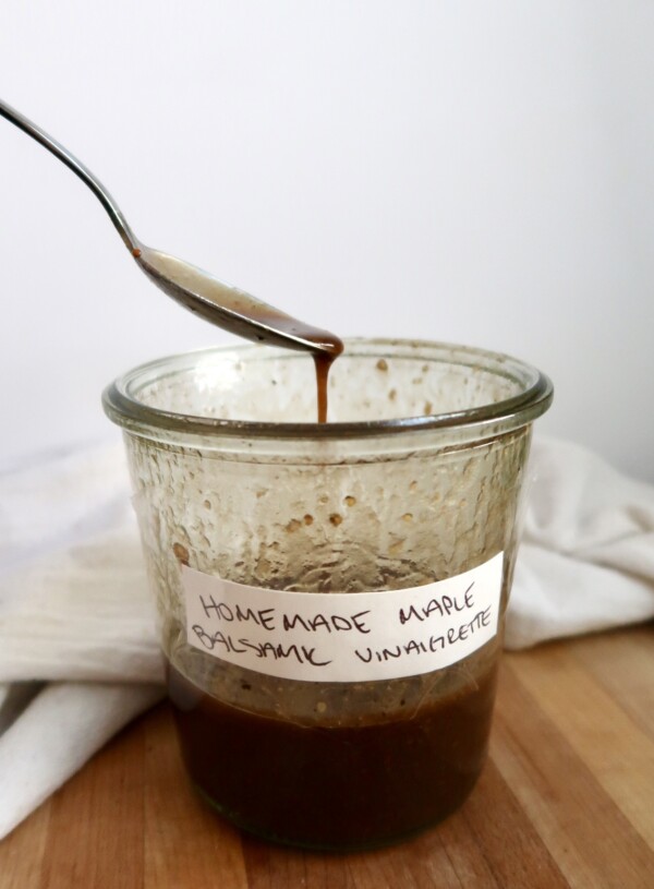 Spoon resting on a glass jar filled with homemade maple balsamic vinaigrette, with a white cloth in the background.