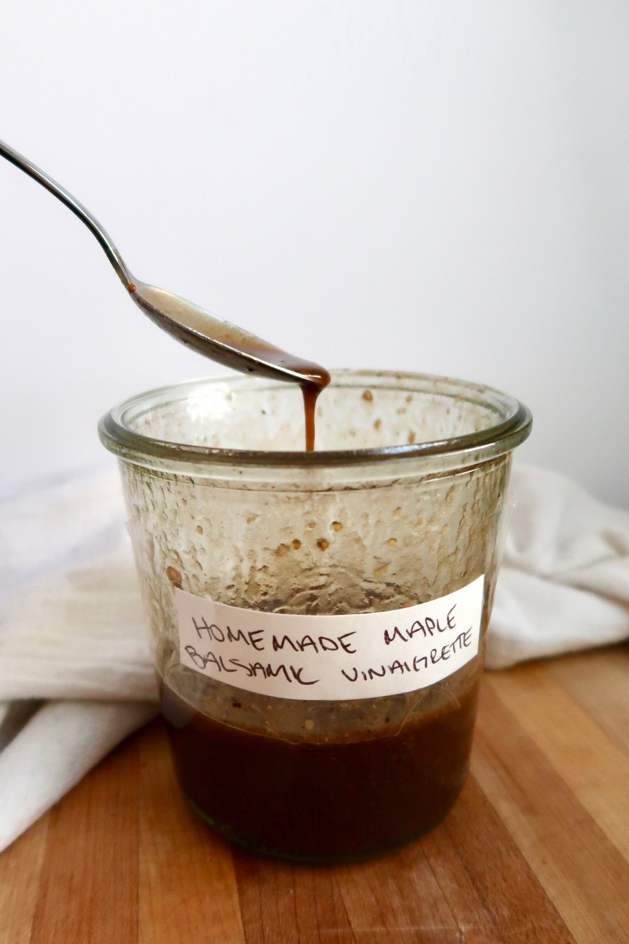 Spoon resting on a glass jar filled with homemade maple balsamic vinaigrette, with a white cloth in the background.