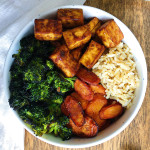 Wooden table with a white bowl with sesame tofu, kale, marinated carrots and brown rice.