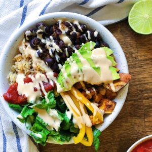 Wooden table with a white bowl of Vegan burrito bowl with spicy lime sauce, with black beans, avocado, rice, and bell pepper with sauce on top and a white and blue napkin in the background.