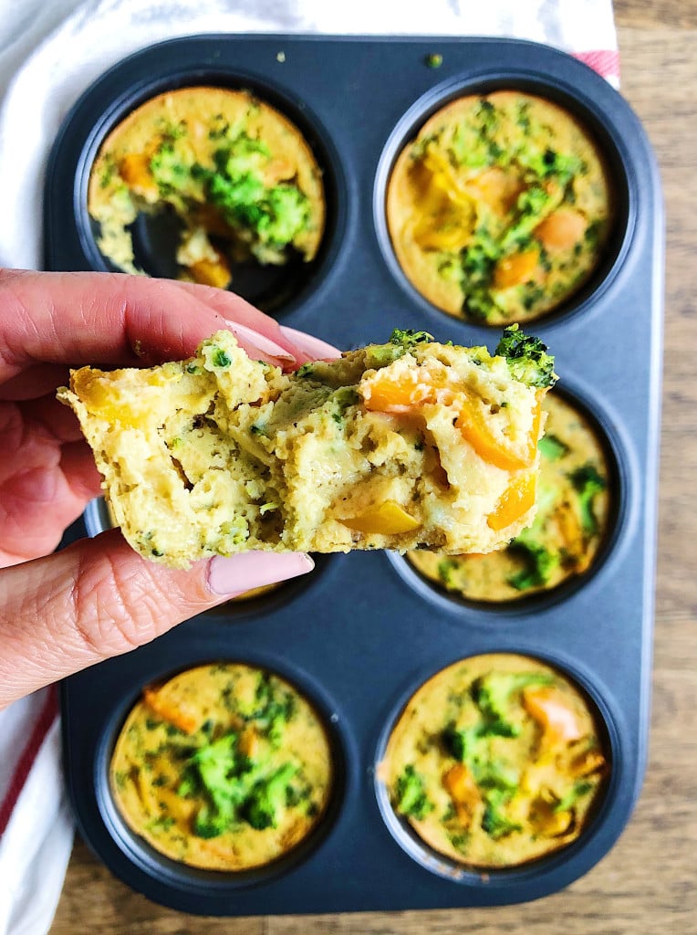 Muffin tin of vegan egg bites with broccoli and orange bell pepper. A hand holding half an egg bite.