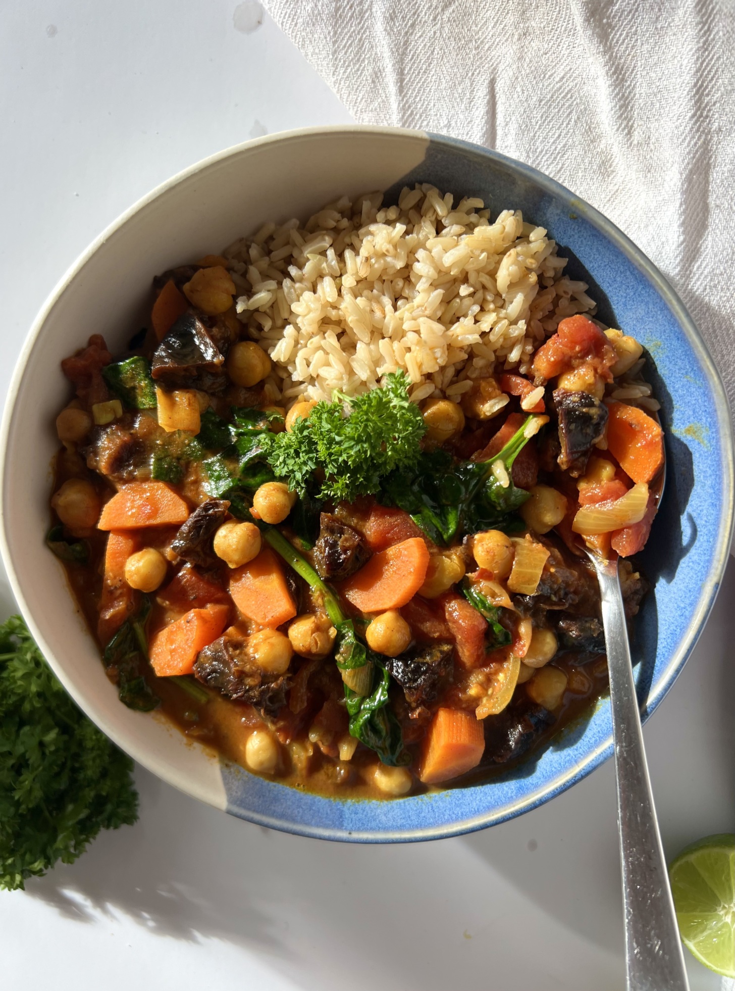 White and blue bowl of spiced chickpea stew with brown rice and parsley for garnish and a spoon in the bowl.