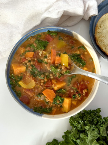 White and blue bowl of sweet potato and kale soup with lentils with bread off to the side and kale as garnish.
