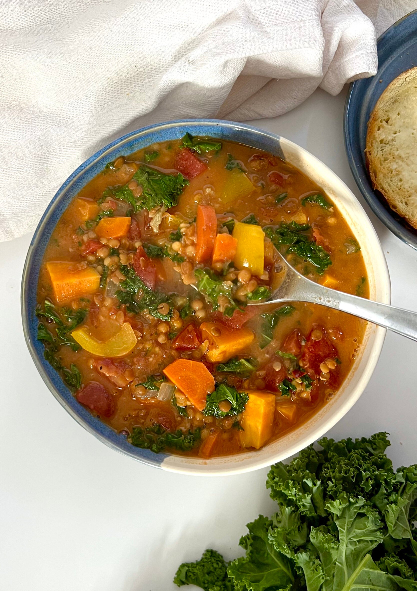 White and blue bowl of sweet potato and kale soup with lentils with a spoon in it and bread off to the side and kale as garnish.