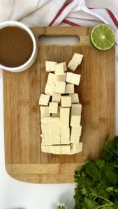 Extra firm tofu on a cutting board cut into 1 inch cubes with teriyaki sauce, lime and cilantro to the side.
