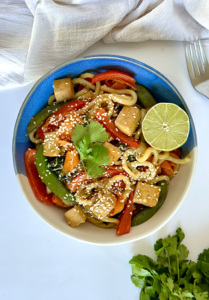 Udon noodle stir fry with tofu and vegetables in it with lime to garnish.