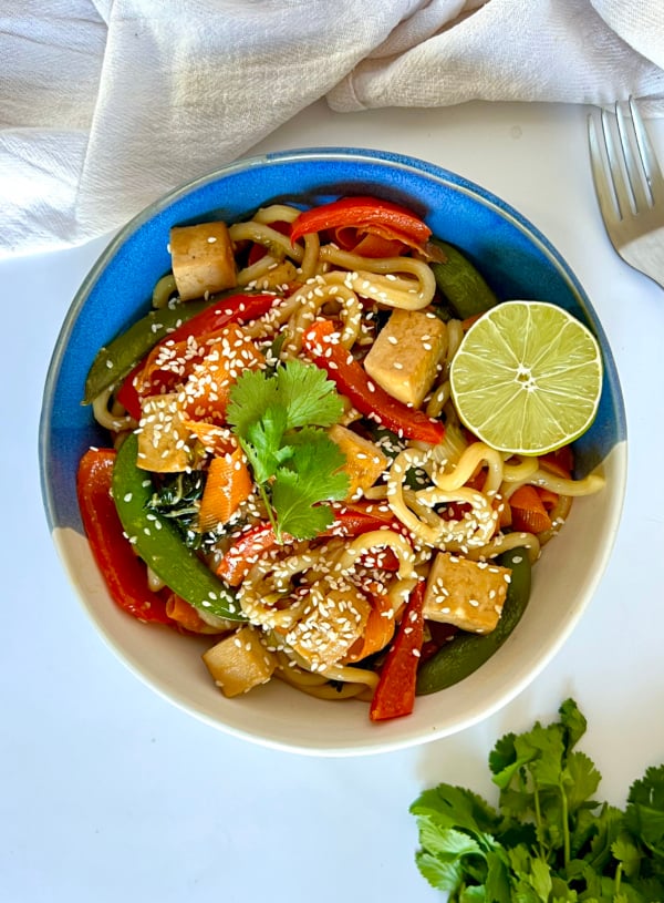 Udon noodle stir fry with tofu and vegetables in it with lime to garnish.