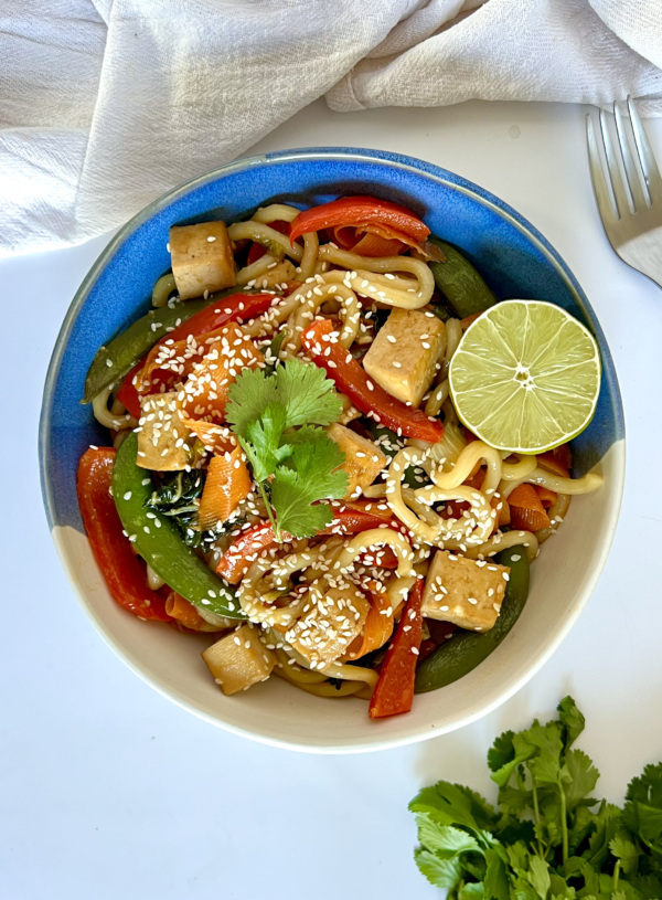 Udon noodle stir fry with tofu and vegetables in it with lime to garnish.