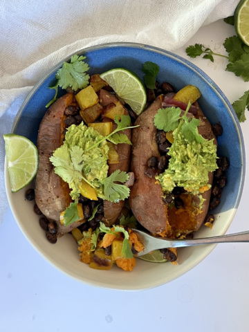 Two baked sweet potatoes in a bowl with black beans, peppers, red onion, mashed avocado, lime and cilantro