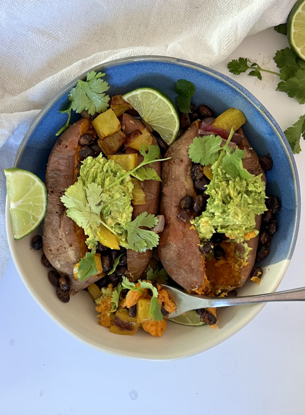 Two baked sweet potatoes in a bowl with black beans, peppers, red onion, mashed avocado, lime and cilantro