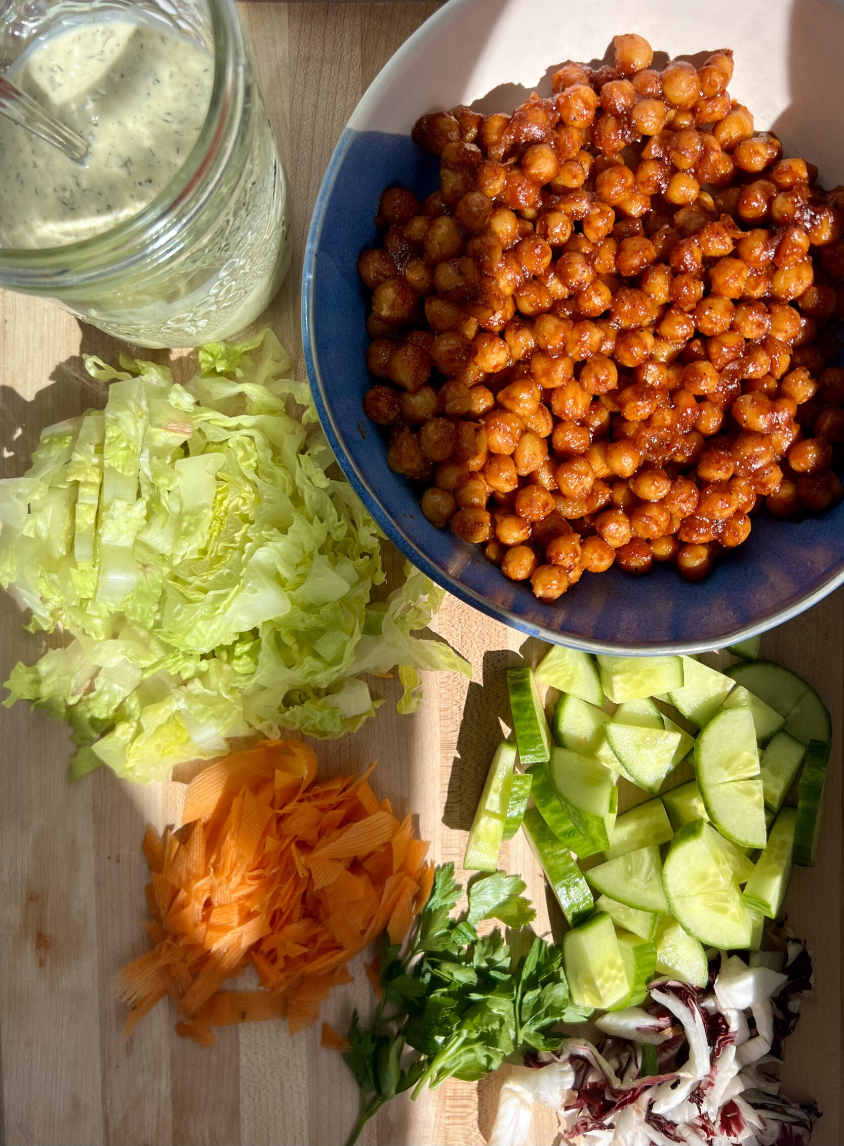 Cutting board with a bowl of chickpeas covered in BBQ sauce, with vegan ranch dressing, and chopped veggies on the side.