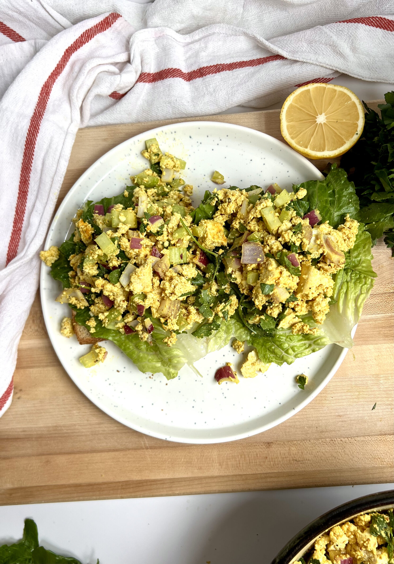 Two pieces of toast with lettuce on top and curry tofu salad, with lemon on the side on top of a cutting board