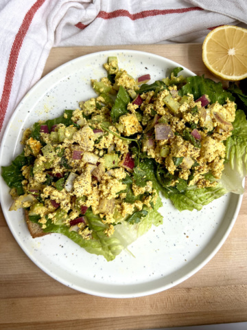 White plate with two pieces of toast on it with lettuce and curry tofu on top with lemon to the side.