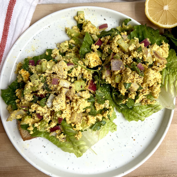 White plate with two pieces of toast on it with lettuce and curry tofu on top with lemon to the side.