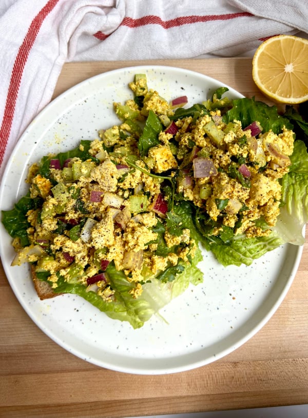 White plate with two pieces of toast on it with lettuce and curry tofu on top with lemon to the side.