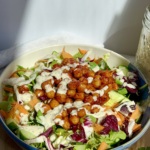 White and blue bowl on a cutting board with a white background and veggies, chickpeas with BBQ sauce, and tahini dressing on top.