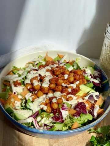 White and blue bowl on a cutting board with a white background and veggies, chickpeas with BBQ sauce, and tahini dressing on top.