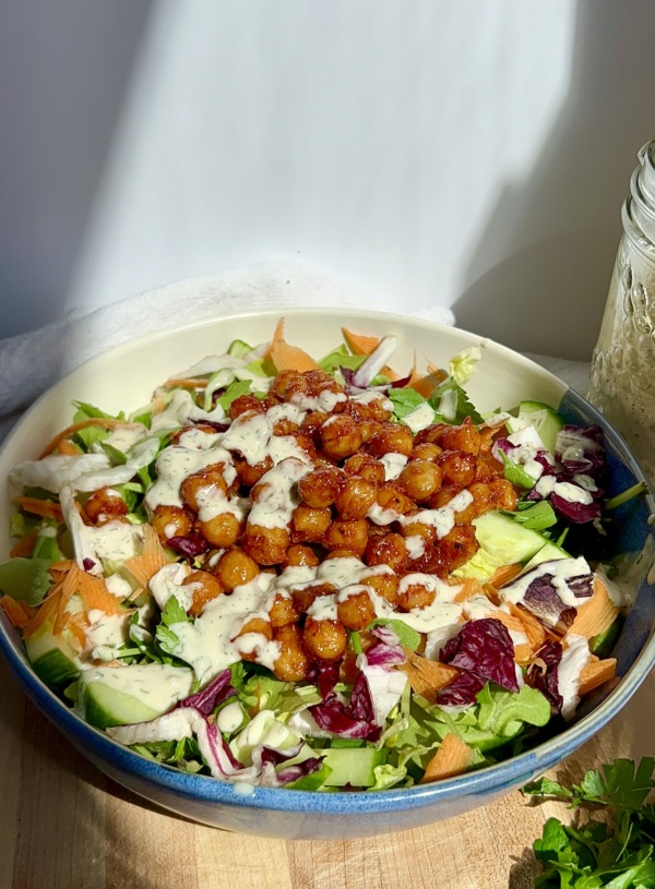 White and blue bowl on a cutting board with a white background and veggies, chickpeas with BBQ sauce, and tahini dressing on top.