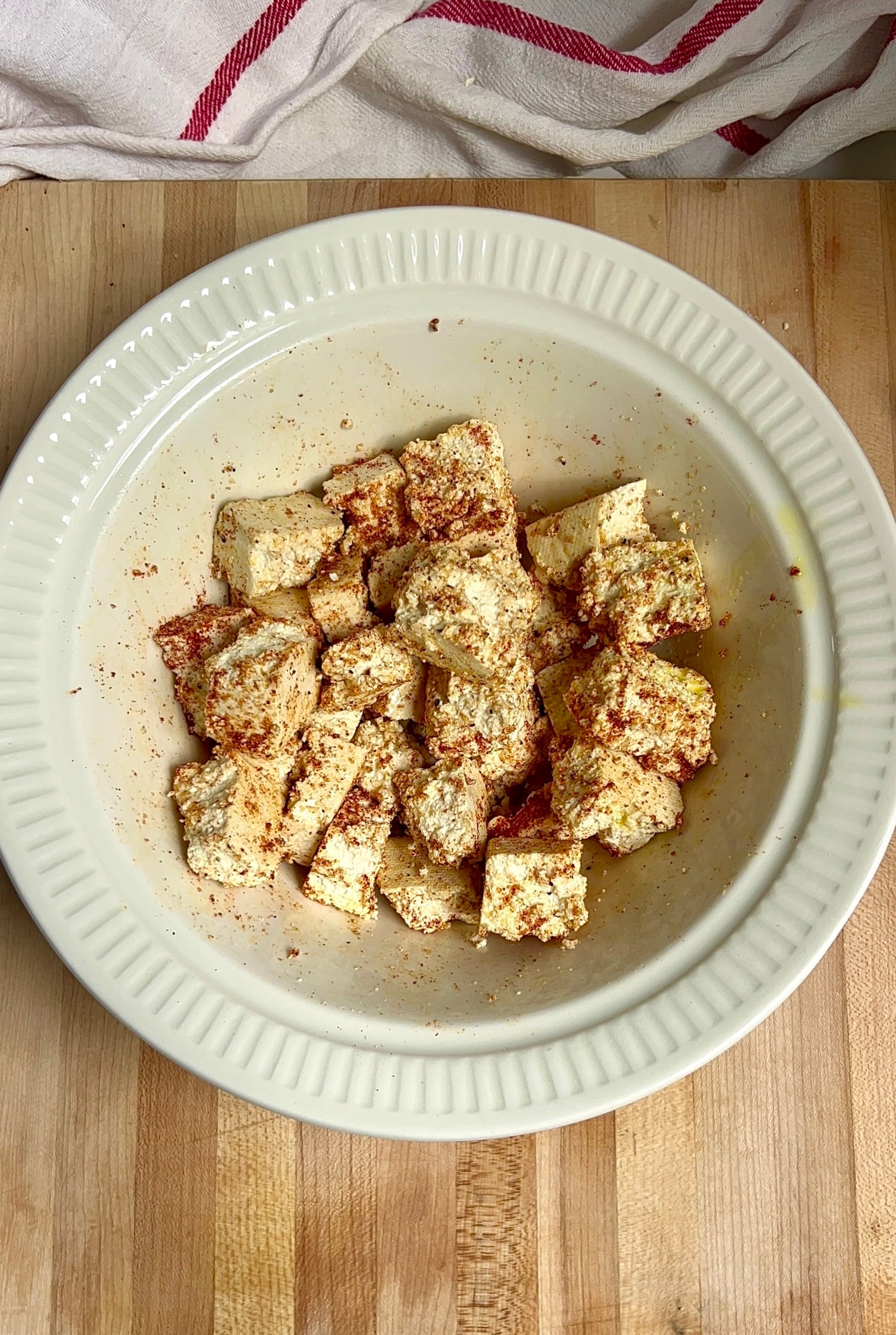 White bowl on a cutting board with tofu ripped into cubes in it with olive oil, garlic powder and smoked paprika tossed together
