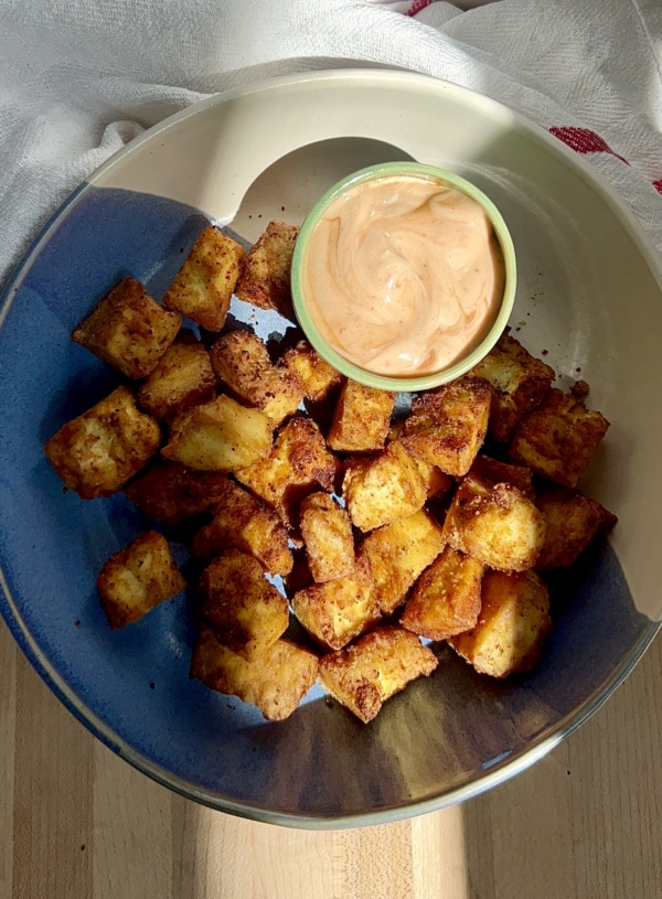 Golden brown tofu in a blue and white bowl on a cutting board with a sauce on the side