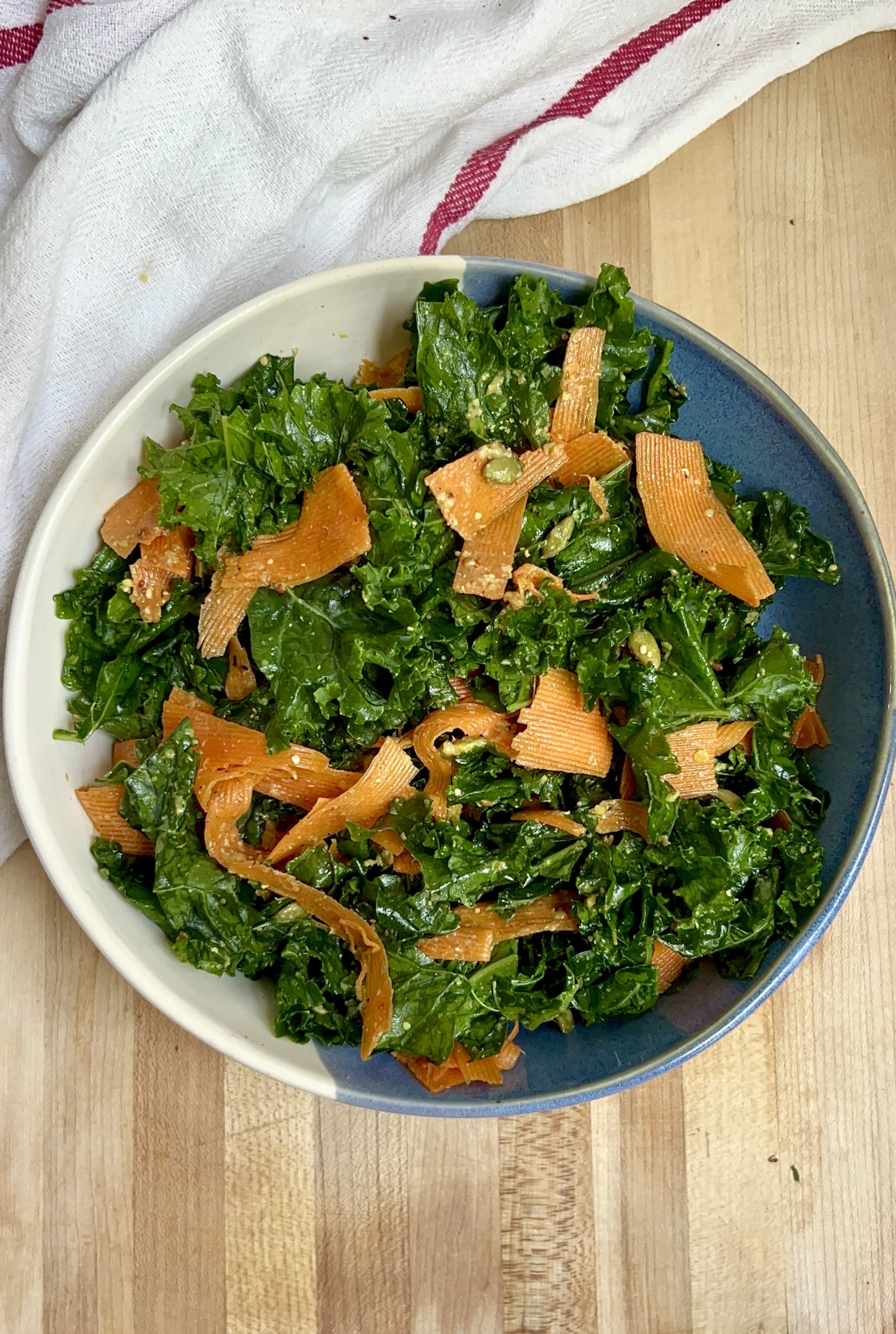 Blue and white bowl of kale salad with grated carrot and pumpkin seeds on top.