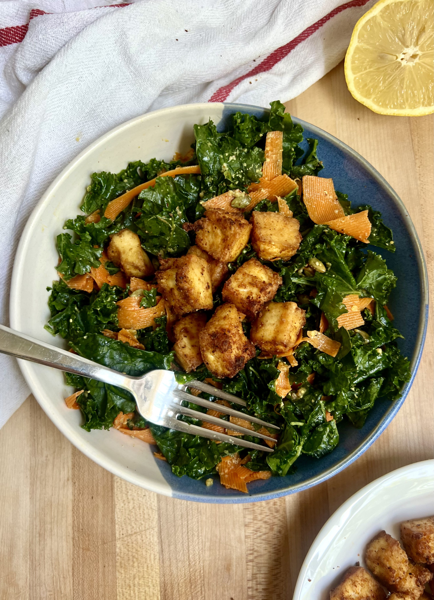 Blue and white bowl with salad and golden brown tofu inside with a lemon off to the side and fork in the bowl
