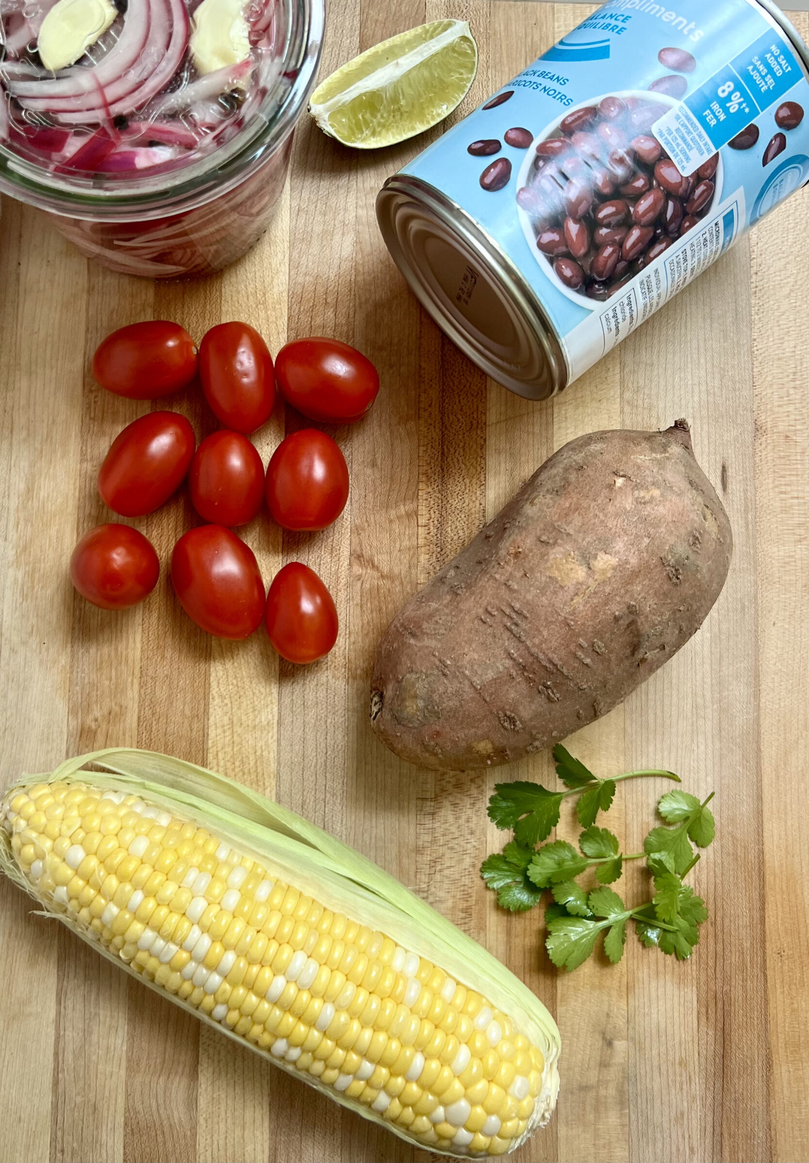Cutting board with pickled red onions in top left corner, black beans, cherry tomatoes, sweet potato, corn and cilantro on it.