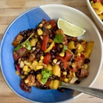 Large bowl with a mixture of peppers, black beans, sweet potato, corn all mixed together and lime on the side and a spoon in it.