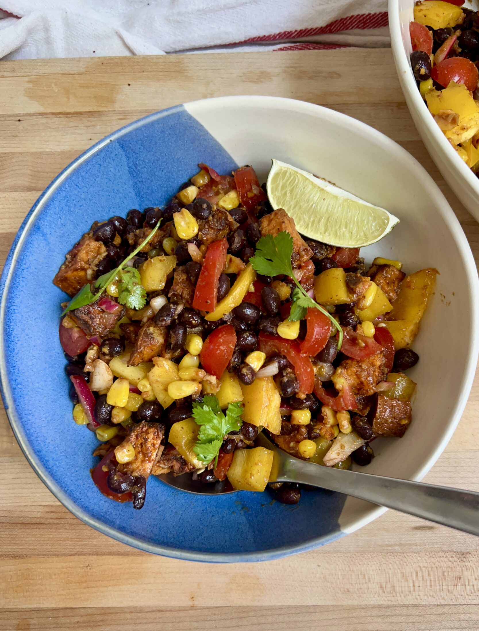 Large bowl with a mixture of peppers, black beans, sweet potato, corn all mixed together and lime on the side and a spoon in it.