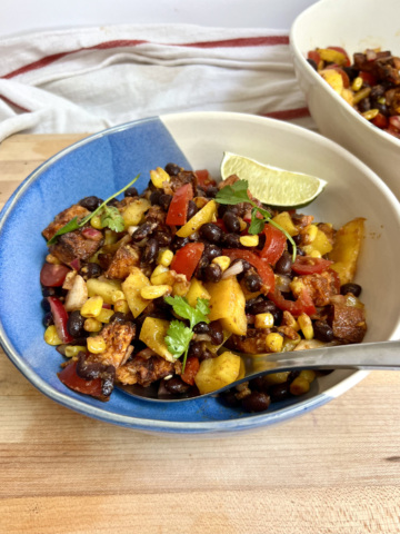 Large bowl with a mixture of peppers, black beans, sweet potato, corn all mixed together and lime on the side and a spoon in it.