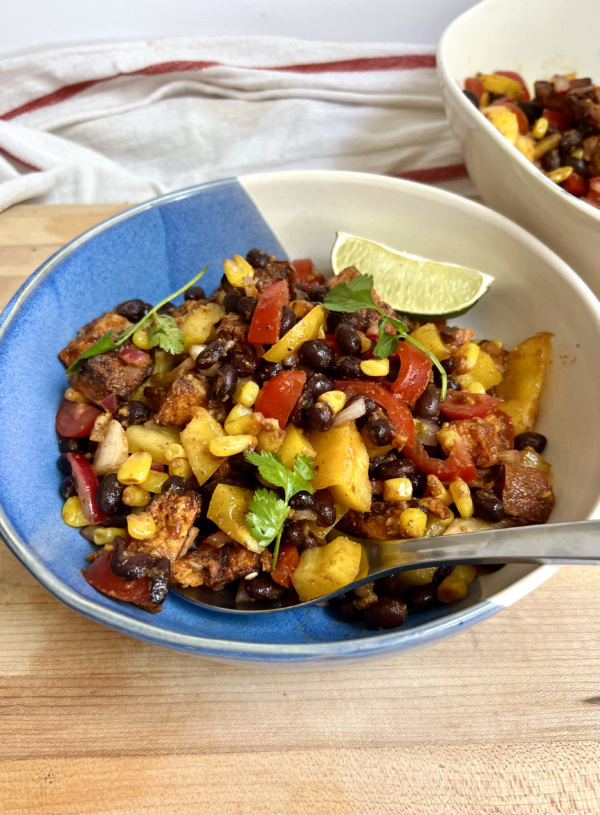 Large bowl with a mixture of peppers, black beans, sweet potato, corn all mixed together and lime on the side and a spoon in it.