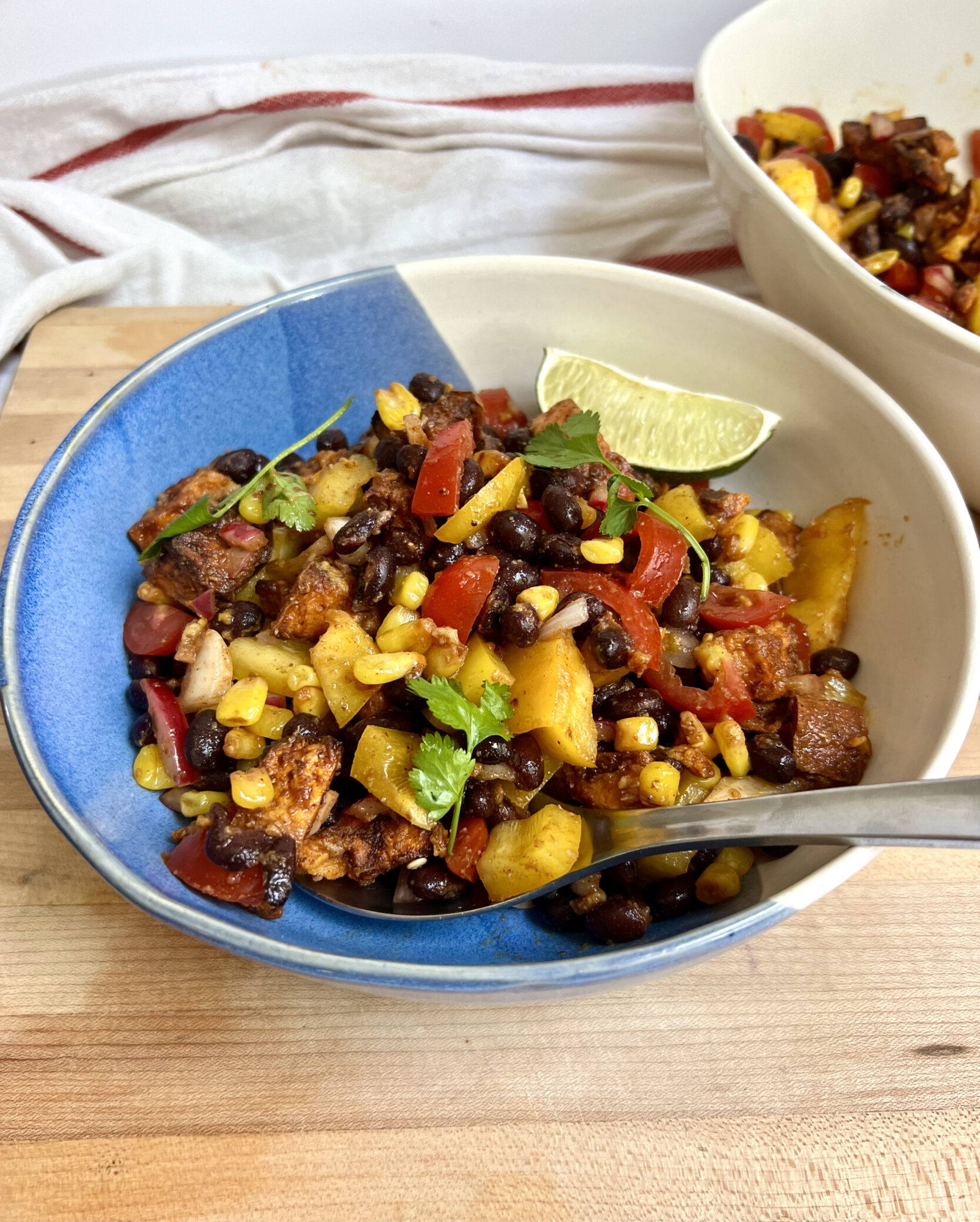 Large bowl with a mixture of peppers, black beans, sweet potato, corn all mixed together and lime on the side and a spoon in it.