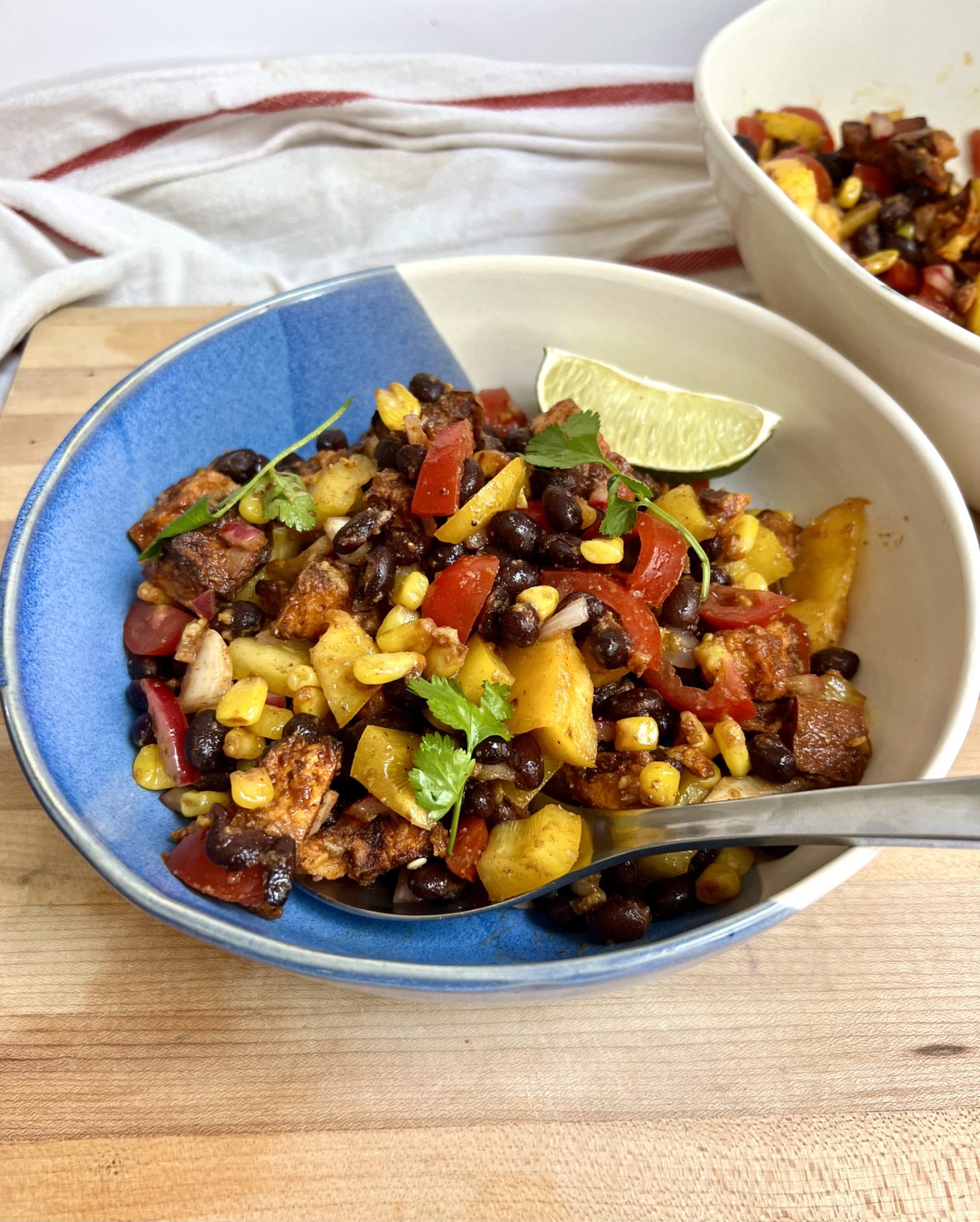 Large bowl with a mixture of peppers, black beans, sweet potato, corn all mixed together and lime on the side and a spoon in it.
