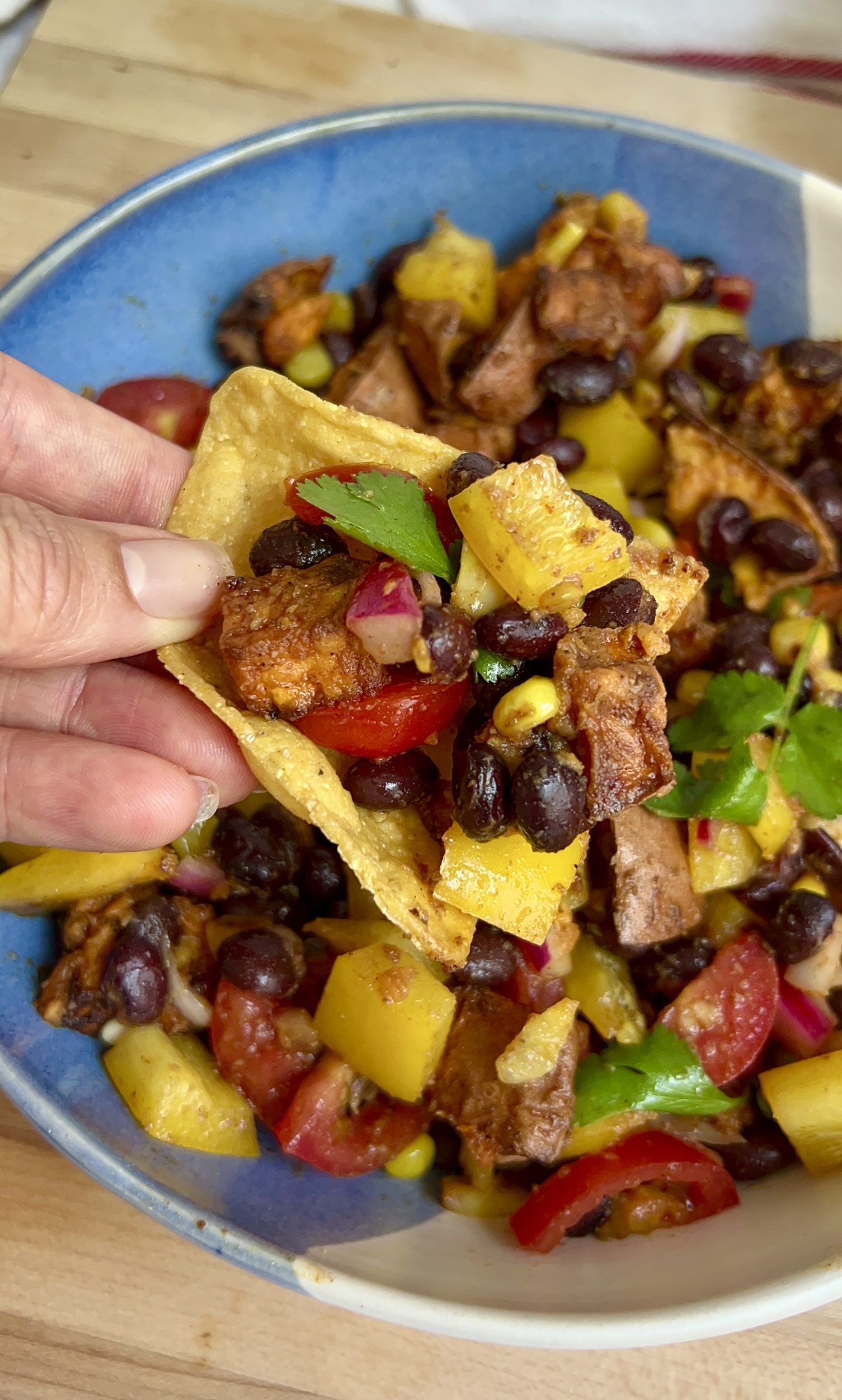 Hand holding a corn ship dipped in a mixture of veggies, black beans and roasted sweet potato. 