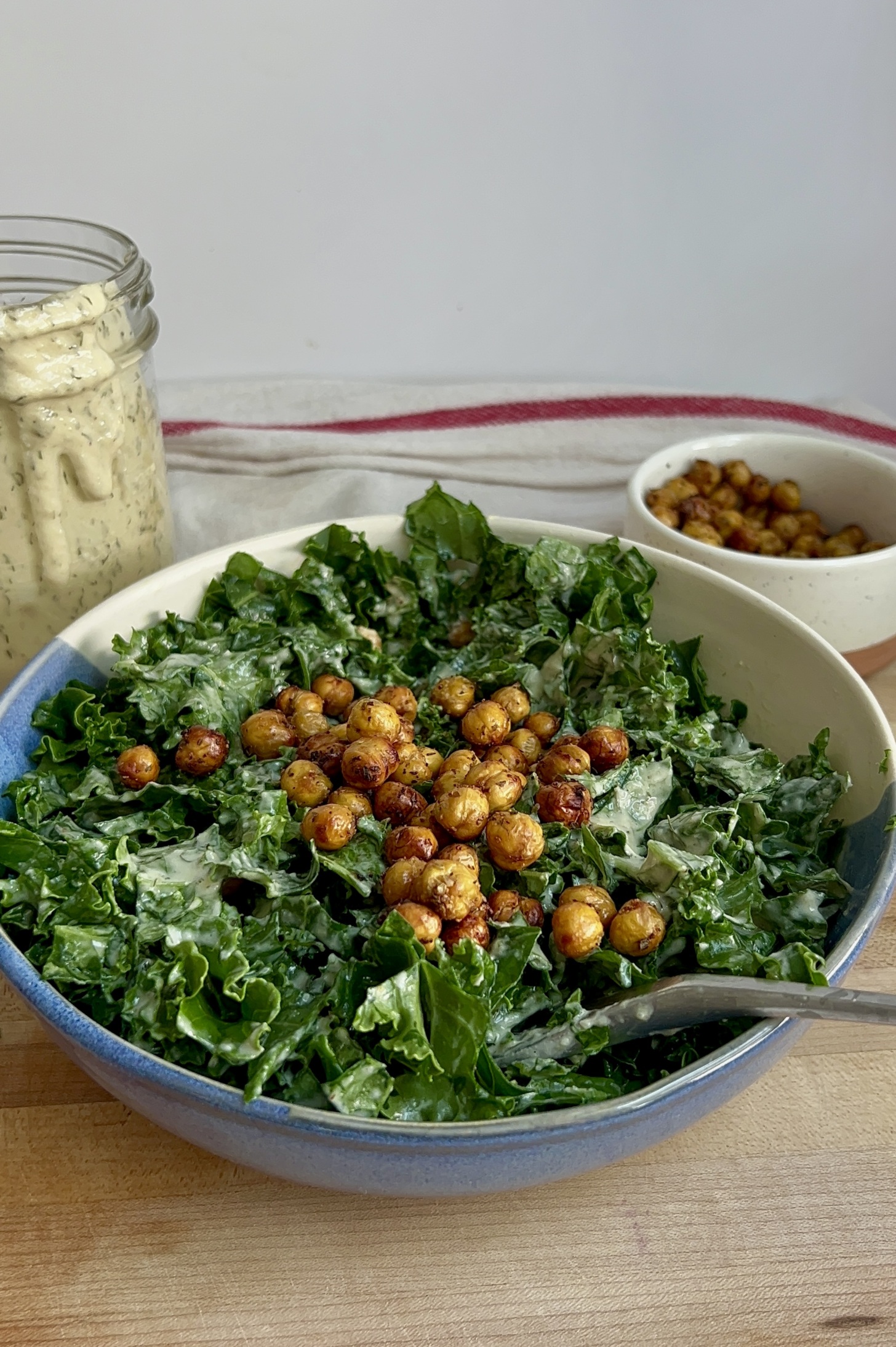 White and blue bowl with massaged kale and tahini dressing and roasted chickpeas on top and a bowl of roasted chickpeas to the side and dressing to the side.