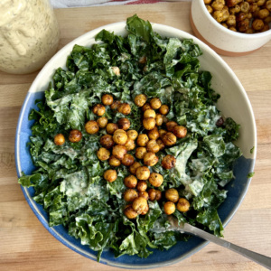 White background with a cutting board and a blue and white bowl with kale with salad dressing inside, roasted chickpeas on top, and salad dressing and a bowl of roasted chickpeas to the side.