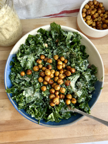 White background with a cutting board and a blue and white bowl with kale with salad dressing inside, roasted chickpeas on top, and salad dressing and a bowl of roasted chickpeas to the side.