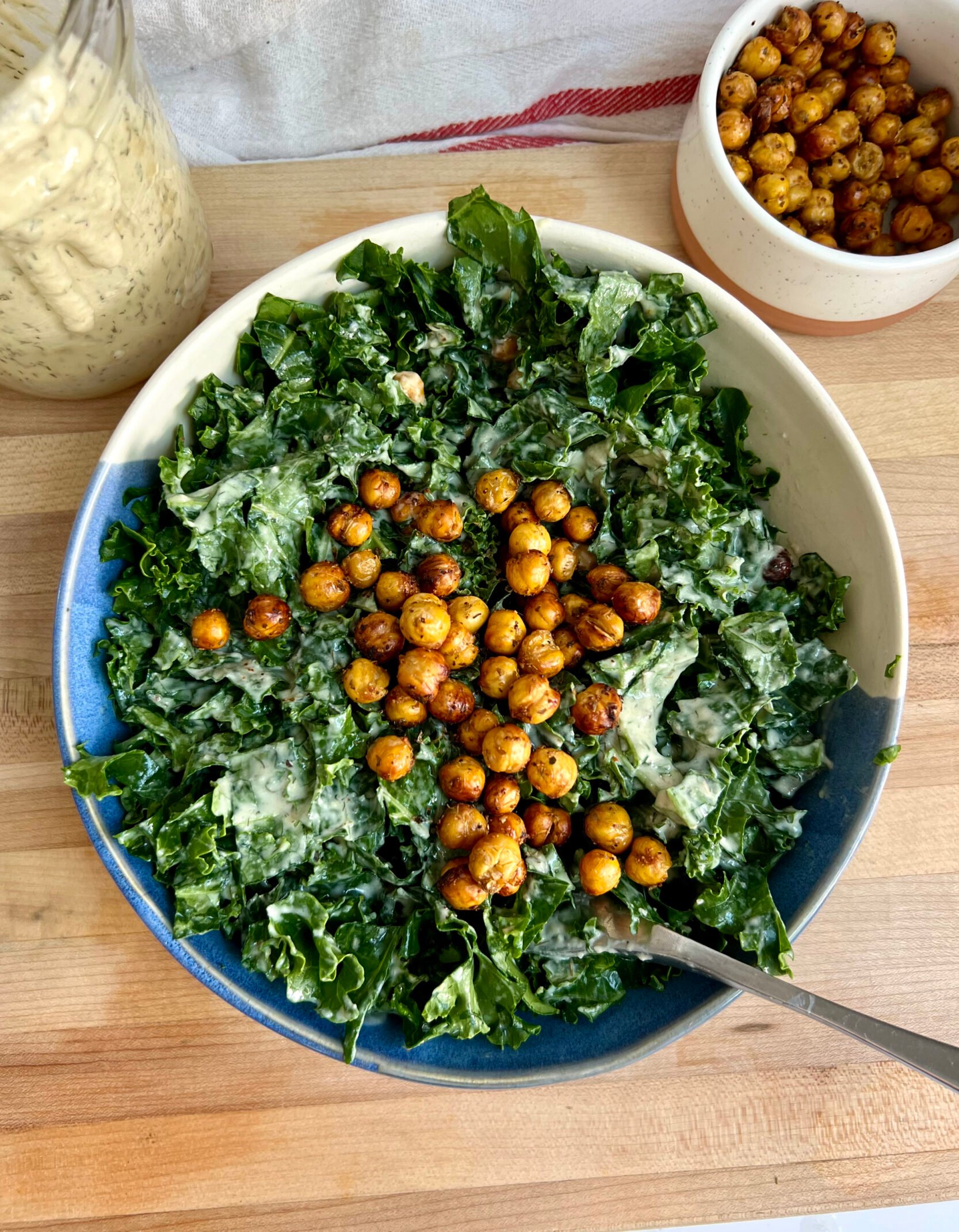 White background with a cutting board and a blue and white bowl with kale with salad dressing inside, roasted chickpeas on top, and salad dressing and a bowl of roasted chickpeas to the side.