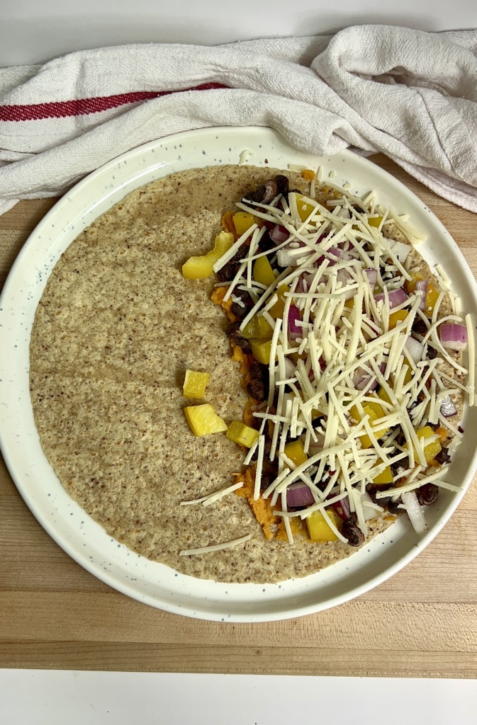 Plate on parchment paper with a wrap on top and mashed sweet potato, bell pepper, red onion, black beans and cheese spread on half.