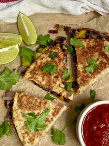 Cooked quesadilla sliced into 3 on parchment paper with cilantro on top, lime to the side and salsa next to it.