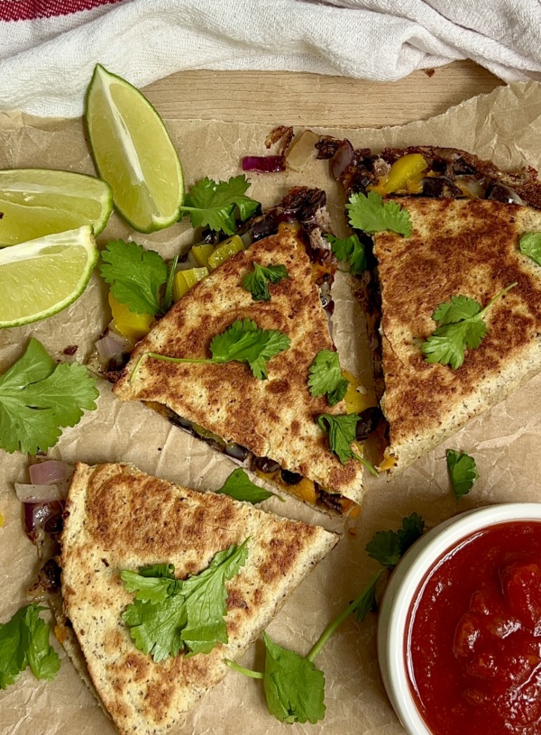 Cooked quesadilla sliced into 3 on parchment paper with cilantro on top, lime to the side and salsa next to it.