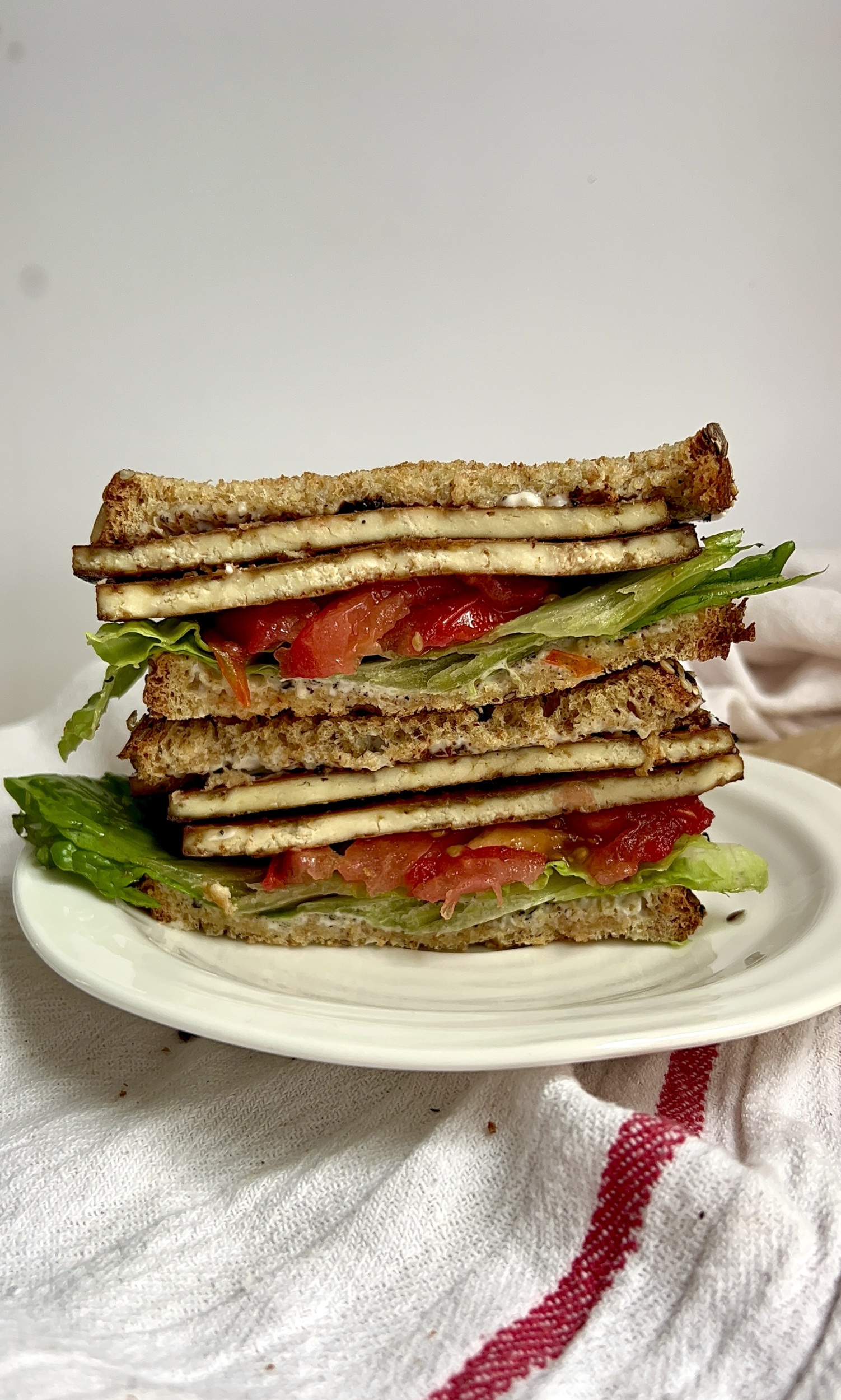 White plate with a sandwich stacked on top with tofu bacon, lettuce and tomato sliced legnthwise.