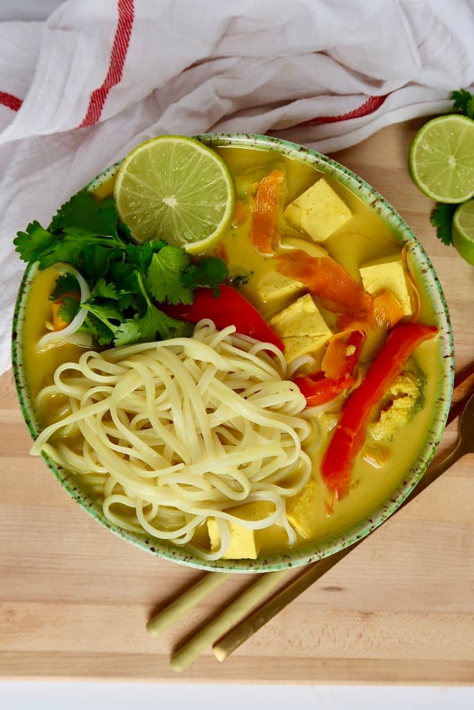 Large bowl with yellow curry broth inside with tofu, noodles, red bell pepper and lime to the side.